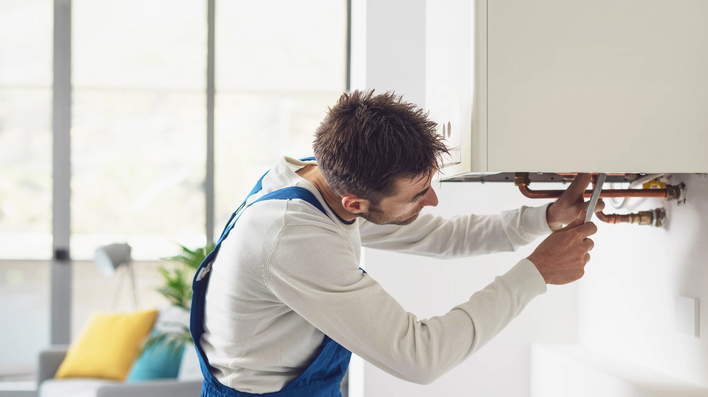 Photographie réaliste d'un artisan plombier professionnel dans un intérieur résidentiel, effectuant un diagnostic ou une intervention sur une tuyauterie/radiateur. L'artisan porte une tenue de travail appropriée (vêtements de métier, outils à la ceinture ou à proximité). La scène doit montrer une action concrète liée au dépannage ou la rénovation sanitaire/chauffage, dans un environnement d'habitation authentique, photographiée sous un éclairage naturel professionnel.