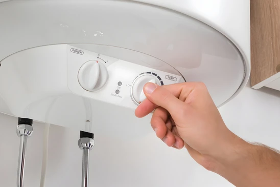 Vue détaillée et rapprochée d'un plombier professionnel effectuant l'installation d'un chauffe-eau électrique ou thermodynamique dans un local technique résidentiel. L'artisan, équipé d'outils appropriés, doit être en train de raccorder les tuyauteries d'arrivée et de distribution au chauffe-eau. Le réservoir doit être clairement visible, de type moderne et professionnel. L'environnement doit montrer un espace technique réel (cave, cellier ou local technique) avec des conduites, des vannes et un travail en cours présenté de manière sérieuse et professionnelle.