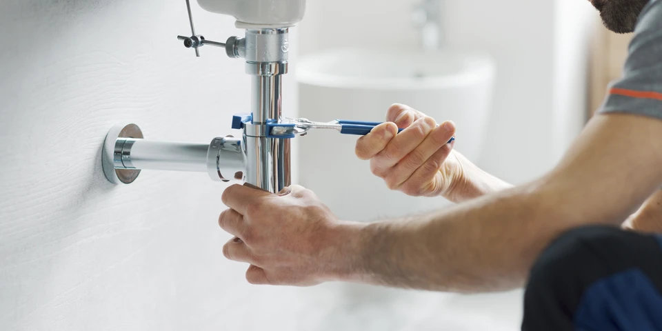 Photographie d'un artisan plombier en tenue professionnelle travaillant à l'intérieur d'une salle de bain en rénovation, montrant à la fois des outils de dépannage urgent (déboucheur, clé anglaise) et les phases finales d'installation sanitaire (raccordement de radiateur, finitions de robinetterie). L'image doit transmettre la polyvalence et la maîtrise technique de l'artisan capable de gérer l'urgence et la rénovation complète en un même lieu.